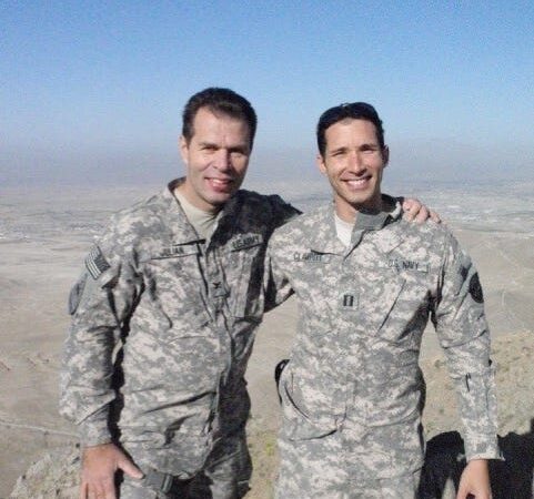 Two Navy soldiers wearing camo gear with their arms around each other standing in the desert and smiling at the camera