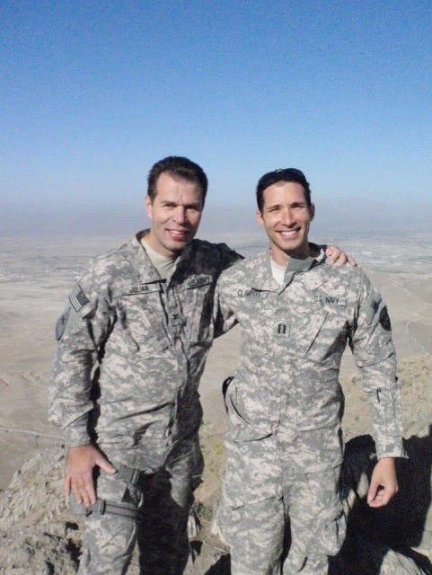 Two Navy soldiers wearing camo gear with their arms around each other standing in the desert and smiling at the camera