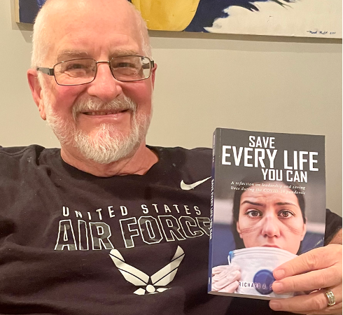 An older bald man with a white beard and glasses wearing a black Air Force t-shirt smiling and sitting on a couch holding up a book titled "Save Every Life You Can"
