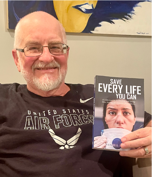 An older bald man with a white beard and glasses wearing a black Air Force t-shirt smiling and sitting on a couch holding up a book titled "Save Every Life You Can"