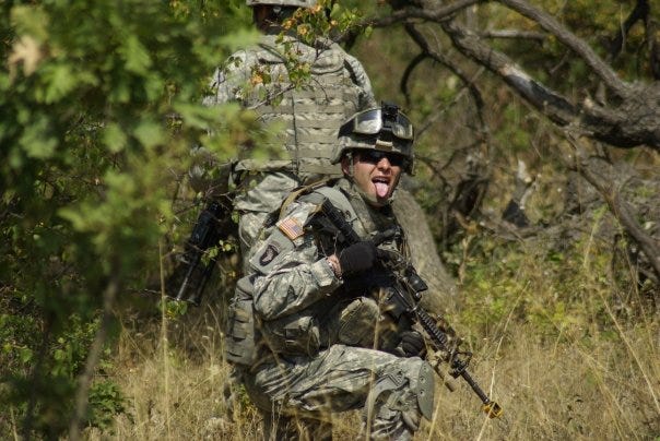 Male American soldier wearing camera crouched with a gun in his hands staring at the camera wearing sunglasses and sticking his tongue out