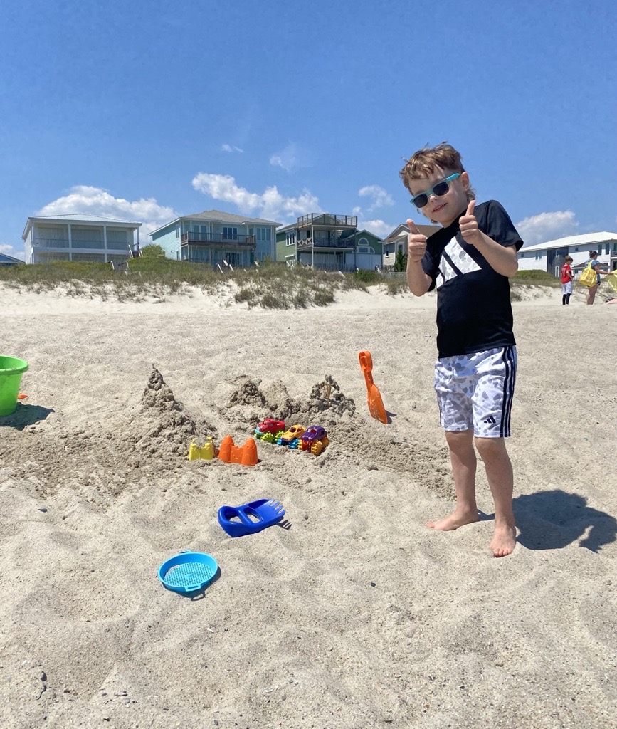 Child giving two thumbs up while standing next to a sand castle on the beach.