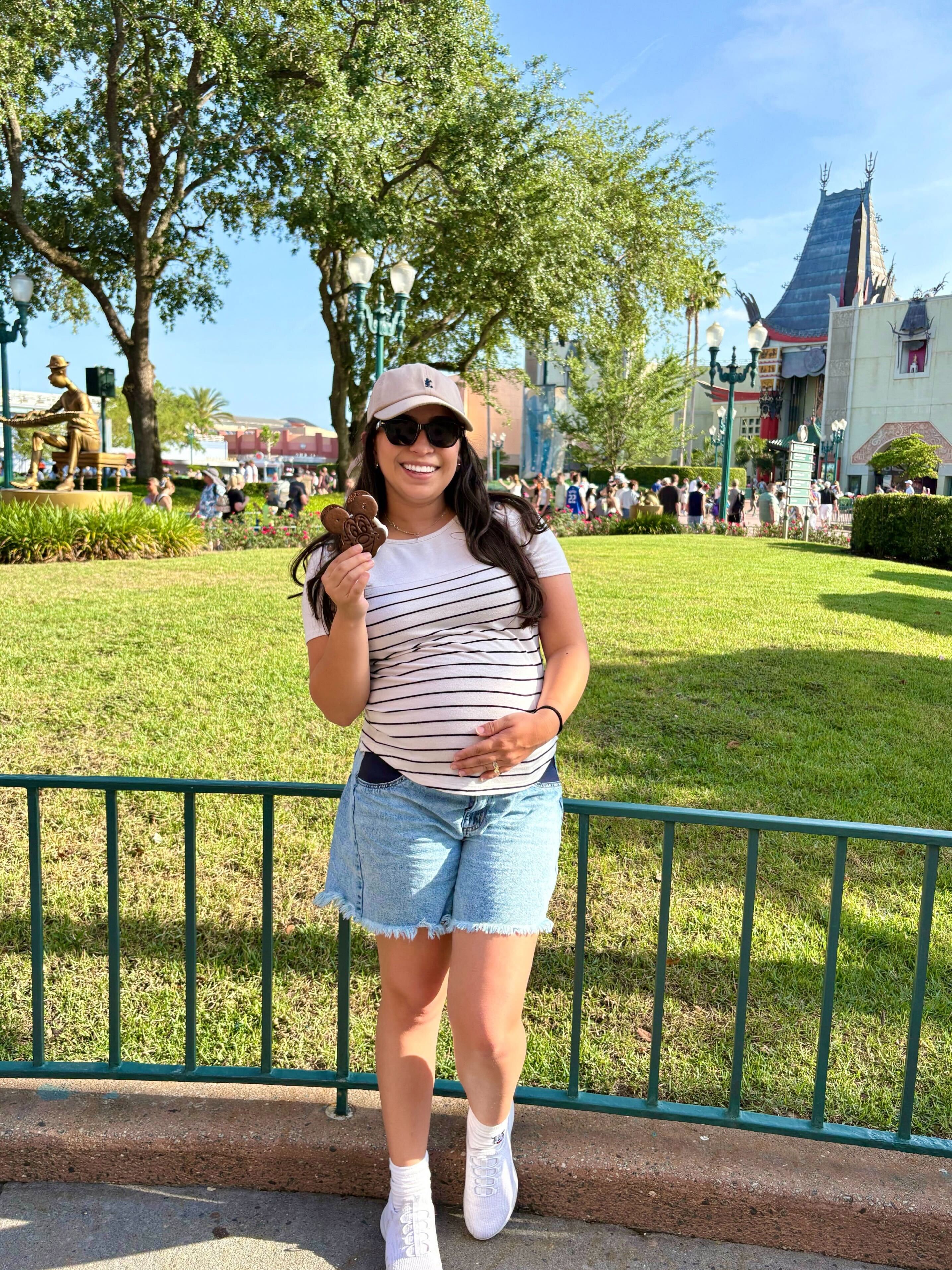 A pregnant woman smiling while holding a Mickey Mouse-shaped ice cream sandwich with a Disney theme park in the background.